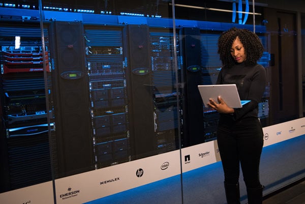 Learning on the job: a woman stands in a data centre while researching on her laptop.